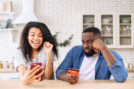 Excited from long awaited purchase multi ethnic woman holds smartphone and rejoices while sad greedy African-American man sitting near with a credit card, not happy to spend moneyの写真素材