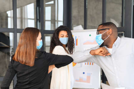 Diverse work team in protective medical masks greet each other with elbows instead of shaking hands standing in contemporary office space. Safety measures for office employmentの写真素材