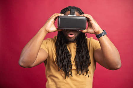 Surprised amazed African American guy with dreadlocks and open mouth isolated over red wall, spending leisure time in virtual reality, playing games in modern VR headset, holding glasses with handsの写真素材