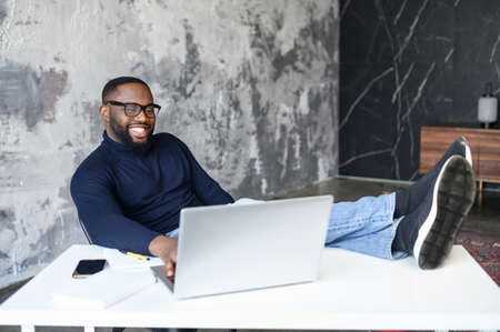 Contemporary African-American male entrepreneur sits in relaxed pose on workplace, put feet on the desk. Successful cheerful black businessman wearing smart casual wear and eyeglasses with a laptopの写真素材