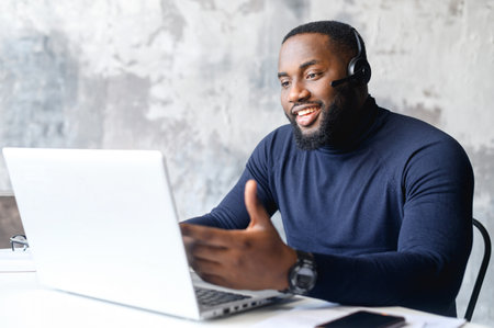 Friendly African-American male entrepreneur wearing wireless headset using a laptop for online communication with customers, a dark skinned employee using hands free device for video conversationの写真素材