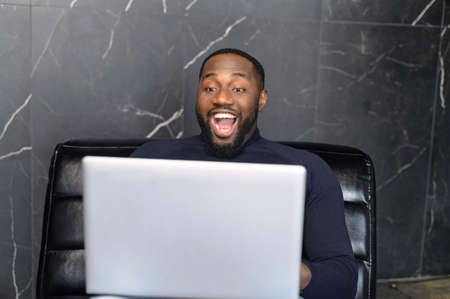 Excited African-American businessman looks at the laptop screen and rejoices, happy black guy scream yes staring with mouth opened at monitor amused with unexpected opportunityの写真素材