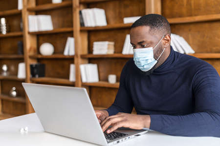 Focused African-American wearing medical mask working with a laptop in contemporary office space with bookshelves on the background, social distancing and protective measures for office employeesの写真素材