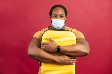 Young African American man in protective face mask holding hugging yellow suitcase, standing isolated on red wall background, feeling stressed because borders closed, unhappy to not be able to travelの写真素材