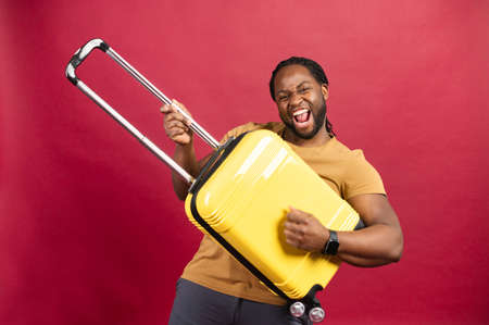 Studio portrait of young African American man with yellow suitcase, playing on it like a guitar, guy on red wall background feeling happy to travel. Summer, holidays, transport, leisure, youth conceptの写真素材