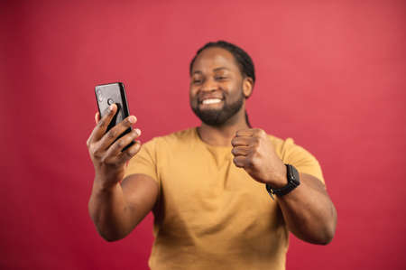 African American ecstatic man biting lip, clenching fist like rejoicing victory or triumph, looking at mobile phone, celebrating success, received great results, got promoted, isolated over red wallの写真素材