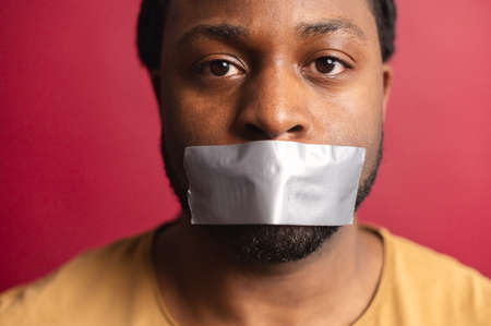 Studio shot of sad African American voiceless speechless guy looking at camera, mouth closed with adhesive tape, suffering from injustice, isolated over red wall background, black lives matter conceptの写真素材