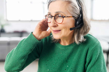 Portrait of a charming elegant senior lady wearing headset and glasses, a mature customer service representative takes a call, close-up portraitの写真素材