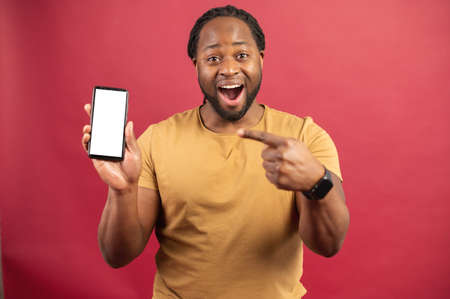 Surprised African-American man holds smartphone in a hand and points finger at empty phone screen, astonishment black guy presenting best deal, advertising new mobile app, isolated on redの写真素材