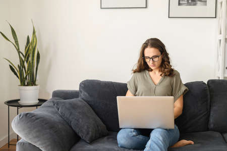 Confident female freelancer using laptop for remote work from home. Focused woman in eyewear typing...