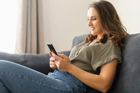 Satisfied young woman with smartphone on the couch rests at home, leaned back in relaxed pose, scrolling news, chatting in social media with friends, messaging online, chatting in date app. Side viewの写真素材