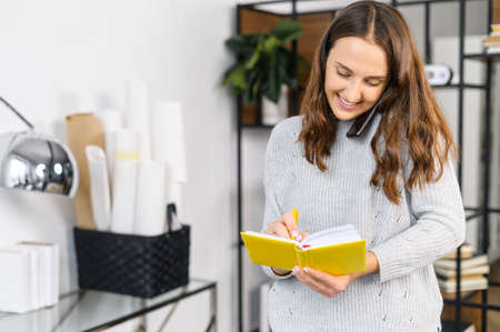 Cheerful woman has phone conversation and writes down into notebook, standing in the office,...