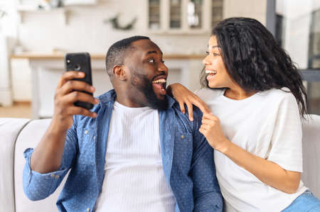 Smiling millennial black couple sit on couch in kitchen have fun browsing Internet on modern smartphone together, happy young husband and wife relax on sofa use cellphone enjoy family weekend at homeの写真素材