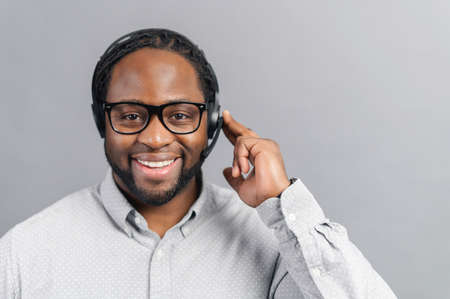 Smiling young African-American office worker in glasses and headset looking at the camera and smiles, positive black man working in the customer service department in touch, isolated on greyの写真素材