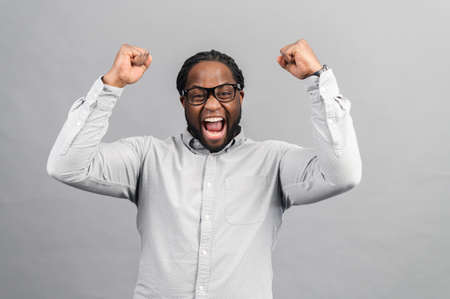 Excited African-American guy wearing smart casual shirt celebrating victory, raising clenched fists up, screams yes happily, black guy yelling ecstatic, triumph concept, studio shot isolated on greyの写真素材