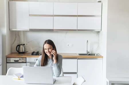 Smiling and friendly young woman talks on the smartphone using laptop computer at home, cheerful female holding phone call speaks with positive face expression, works remotely sitting in the kitchenの写真素材