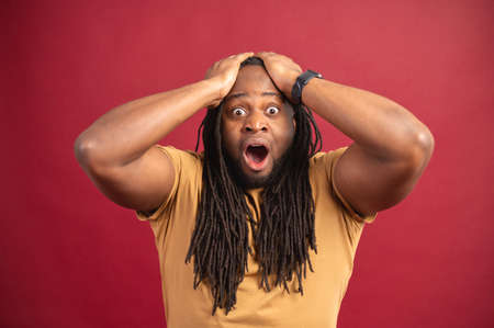 Surprised open-eyed african man with a long dreadlocks staring at the camera with opened mouth, astonished multiracial black guy holding head with both hands isolated on redの写真素材