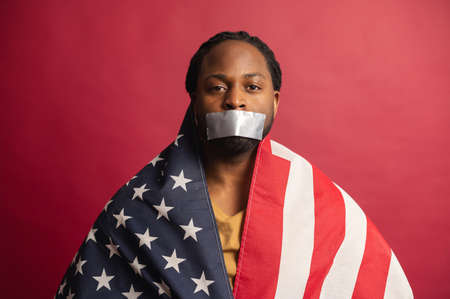 Confident African American fighter for rights voiceless speechless man covered with USA flag, mouth closed with adhesive tape, suffering from injustice, isolated on red wall, black lives matterの写真素材