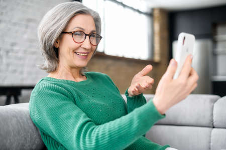 Mature female holding smartphone and talking online on the distance. Senior female using mobile app...
