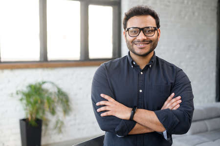 Headshot of skilled hindu male employee standing with arms crossed in modern office, successful confident mixed-race man wearing eyeglasses and smart casual, business portrait of indian entrepreneurの写真素材