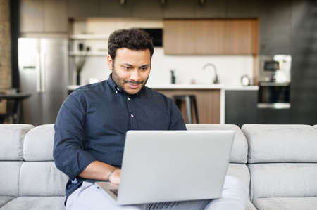 Smiling cheerful mixed-race freelancer guy is using laptop computer sitting on the couch at home,...
