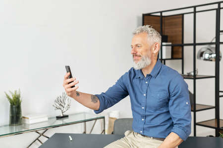 Mature grey haired businessman in smart casual wear is using smartphone for video connection sitting...