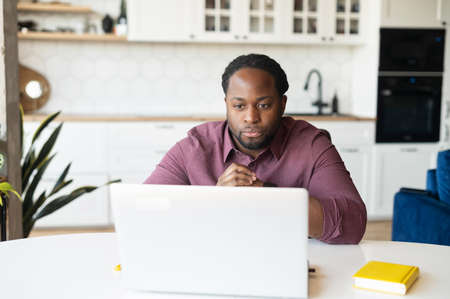 Concentrated African-American guy with dreadlocks staring at computer monitor sitting at the desk in home office, focused black man solving business tasks working with laptop remotely from homeの写真素材