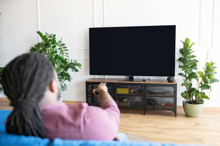 Back view African-American guy with dreadlocks holds remote controller and switching tv channels, watching TV sitting on the couch at home, the lcd TV with empty black screen, copy space, mockupの写真素材
