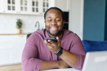 Cheerful African-American man with locks hair sending voice message sitting at home office, positive black guy in casual shirt using voice recognition app, recording audio reportの写真素材