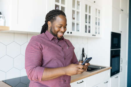 Hilarious guy with dreadlocks using smartphone for communication and messaging online, African-American man enjoys chatting in social networks, looks at the screen and smiles standing in the kitchenの写真素材