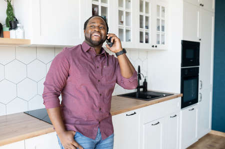 Hilarious black guy with dreadlocks in smart casual shirt enjoys phone conversation with friend or girlfriend, joyful African-American man laughs during pleasant phone conversation standing in kitchenの写真素材