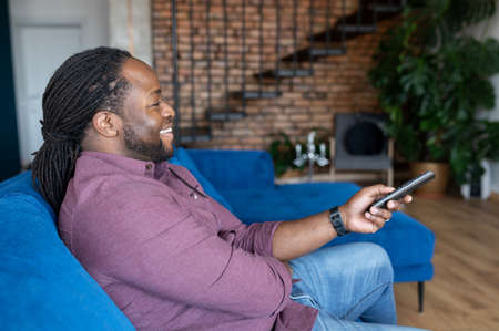 Cheerful and happy African-American man with dreadlocks holding TV remote controller, black guy watching television shows, enjoying favorite series, resting on the couch at home, weekend leisureの写真素材