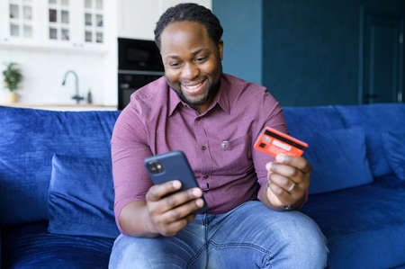 African-American man paying online, makes transaction on his bank account, holds smartphone and...