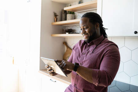 Happy and cheerful African-American guy scrolling and typing on touchscreen of digital tablet, using new educational app or playing video game in the kitchen at home, enjoying chatting onlineの写真素材