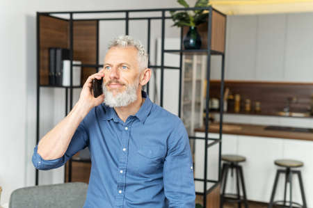 Contemporary middle aged businessman in smart casual wear talking on the smartphone and looks at the camera with a toothy smile, mature blue eyed handsome man has phone conversation indoorの写真素材