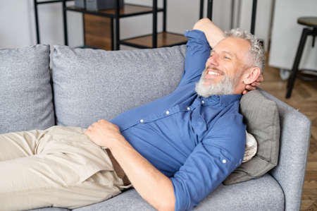 Serene grey-haired hipster guy rests on the couch in cozy living room, mature man lying down on the sofa, relaxing at home, smiling senior male reflecting, daydreaming on the sofa, has restful momentの写真素材
