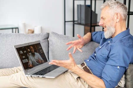 Mature bearded man talking online with African-American male colleague or friend via video...