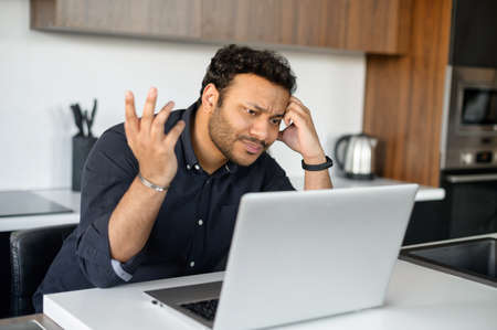 Confused indian man in smart casual shirt looking at the laptop screen, misunderstanding what...