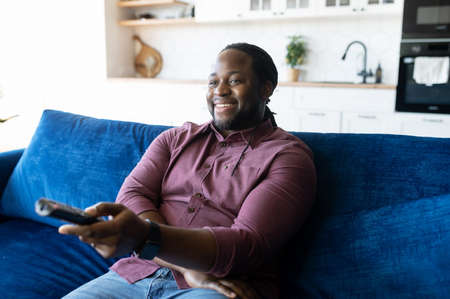 Smiling African-American man with dreadlocks holding TV remote controller, black guy watching television shows, enjoying favorite series, resting on the couch at home, weekend leisureの写真素材