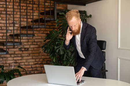 Busy and responsible red-haired bearded modern businessman in formal suit talking on the smartphone...