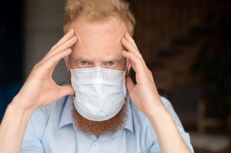 Concerned redhead man wearing protective medical mask staring at the camera with fear in his eyes, guy in doubt holding on to temples, taking measures to protect against covid, worry about futureの写真素材