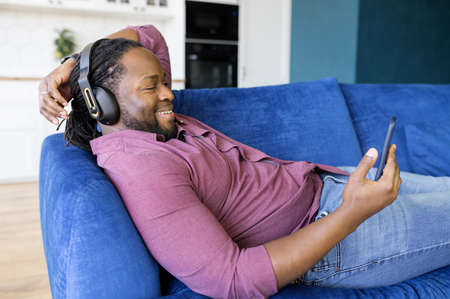 Happy and relaxed African-American man in headphones spends leisure time watching movies on the smartphone, black guy in earphones lying down on the sofa and using phone, listening musicの写真素材