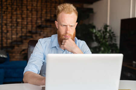 Absorbed and thoughtful red haired businessman staring at the computer monitor, concentrated ginger head freelancer using laptop in the home office, solving tasks, browse internet, researching topicの写真素材