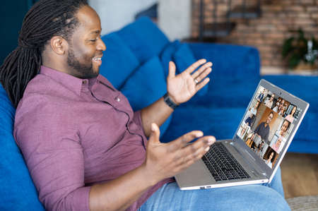 Happy African-American guy makes video call on the laptop, talking and discussing with group of diverse colleagues on the distance, using new computer app for video connection, takes part in webinarの写真素材