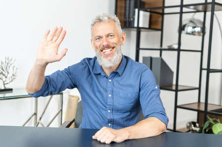 Cheerful middle-aged male employee in smart casual shirt looks at the camera and waving hi, greeting...