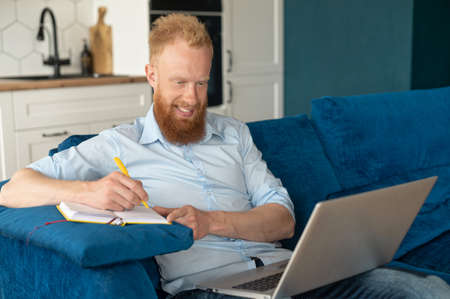 Smiling young redhead man using laptop computer for studying and work on the distance, bearded...