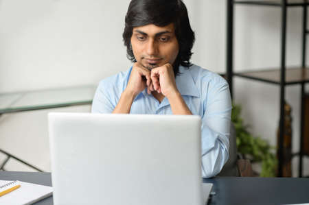 Focused clever indian man in smart casual shirt using laptop indoor, assembled hindu male office...