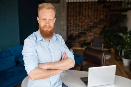 Successful redhead man wearing smart casual shirt stands in confident pose with arms crossed, confident red haired male entrepreneur in home office looks at the cameraの写真素材