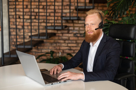 Inspired smiling young redhead businessman using wireless headset and laptop computer for online communication on the distance, cheerful optimistic businessman wearing headphones with mic looks awayの写真素材