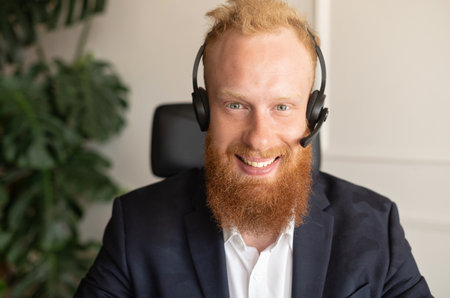 Headshot of smiling young redhead man using wireless headset for online communication on the distance, cheerful optimistic businessman wearing headphones with mic looks at camera, close-up portraitの写真素材